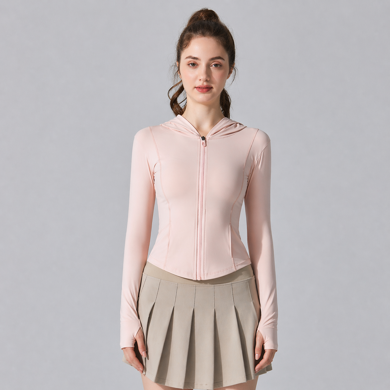Woman wearing a light pink zip-up hoodie and beige pleated skirt on a gray background