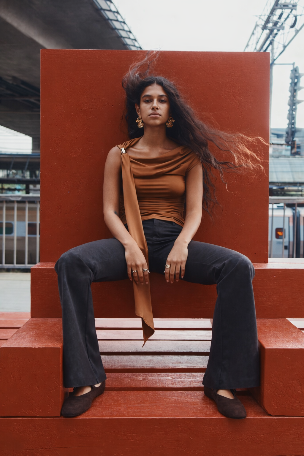 Woman sitting on red steps wearing a brown top and black pants, with a blurred urban background.