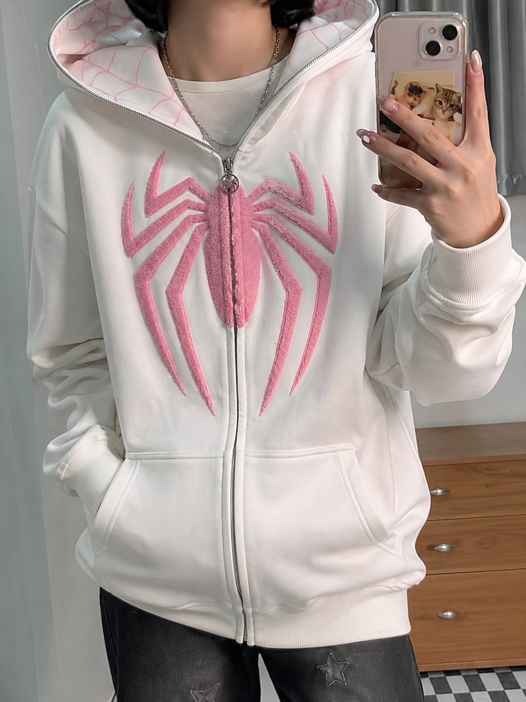 Person wearing a white hoodie with a pink spider emblem, taking a mirror selfie.
