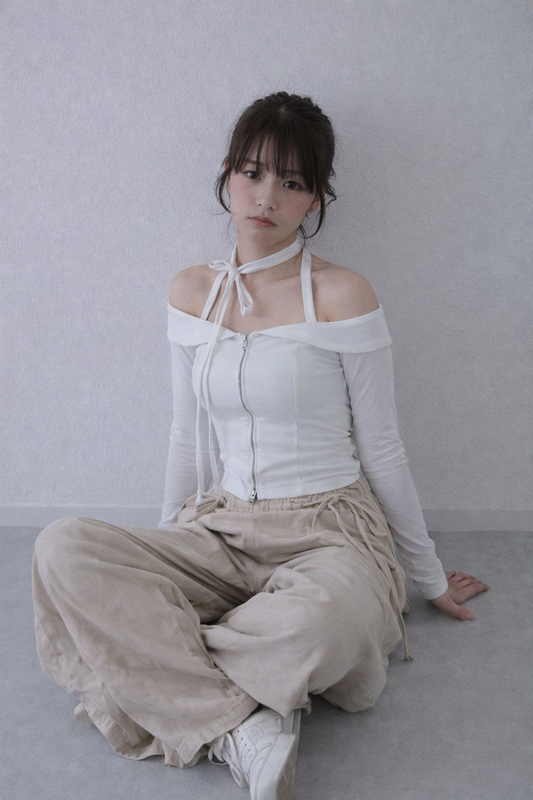 Woman wearing a white off-shoulder top and beige pants sitting on a gray floor against a light gray wall.