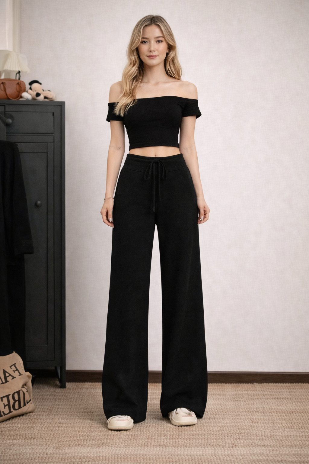 Woman wearing a black off-shoulder top and wide-leg pants standing in a room with a black cabinet and beige rug.