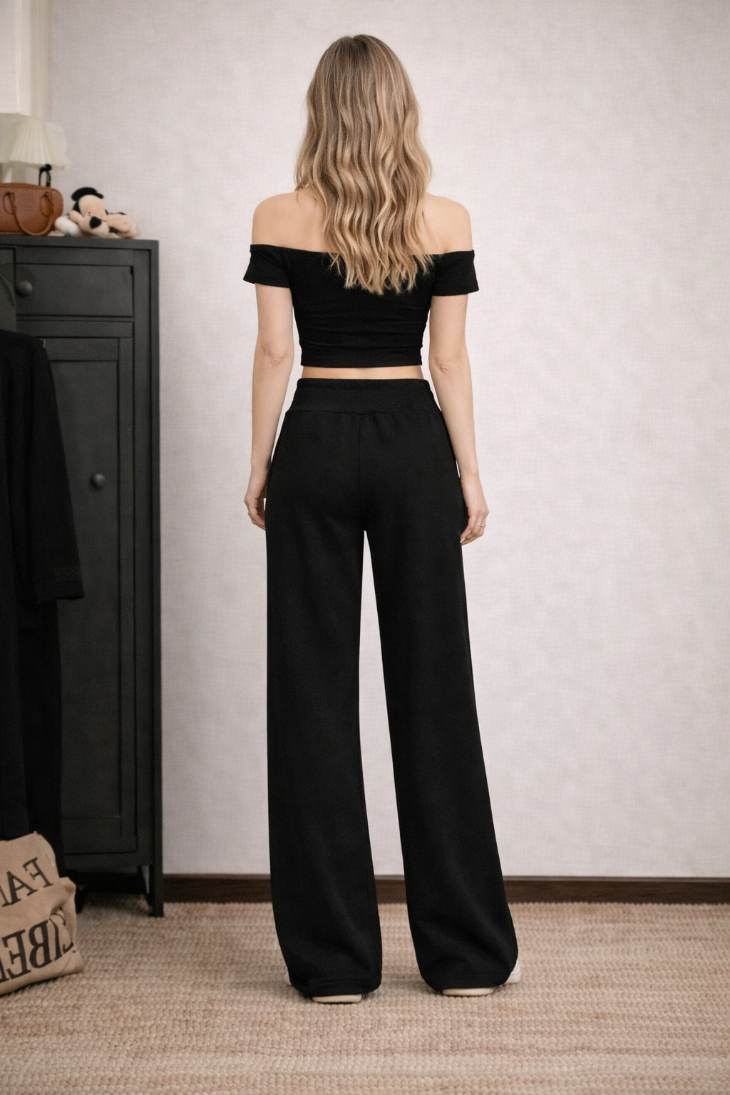 Woman wearing a black off-shoulder top and wide-leg pants in a room with a dresser and bag.