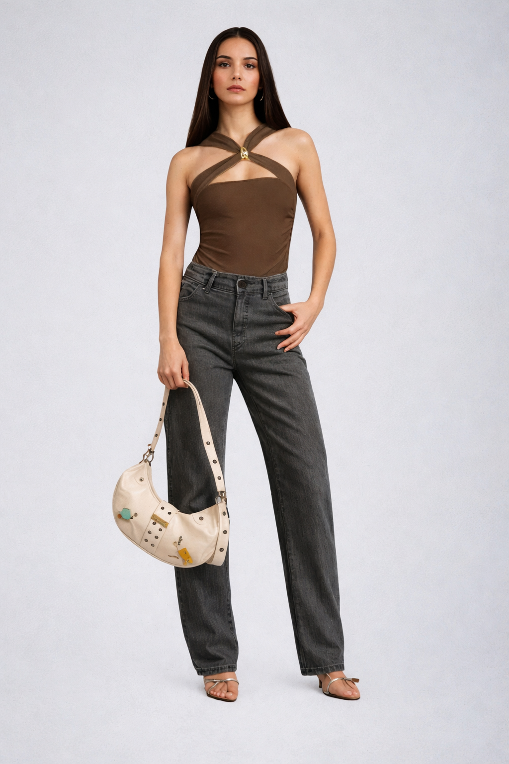 Woman wearing a brown top and gray jeans holding a beige handbag on a white background