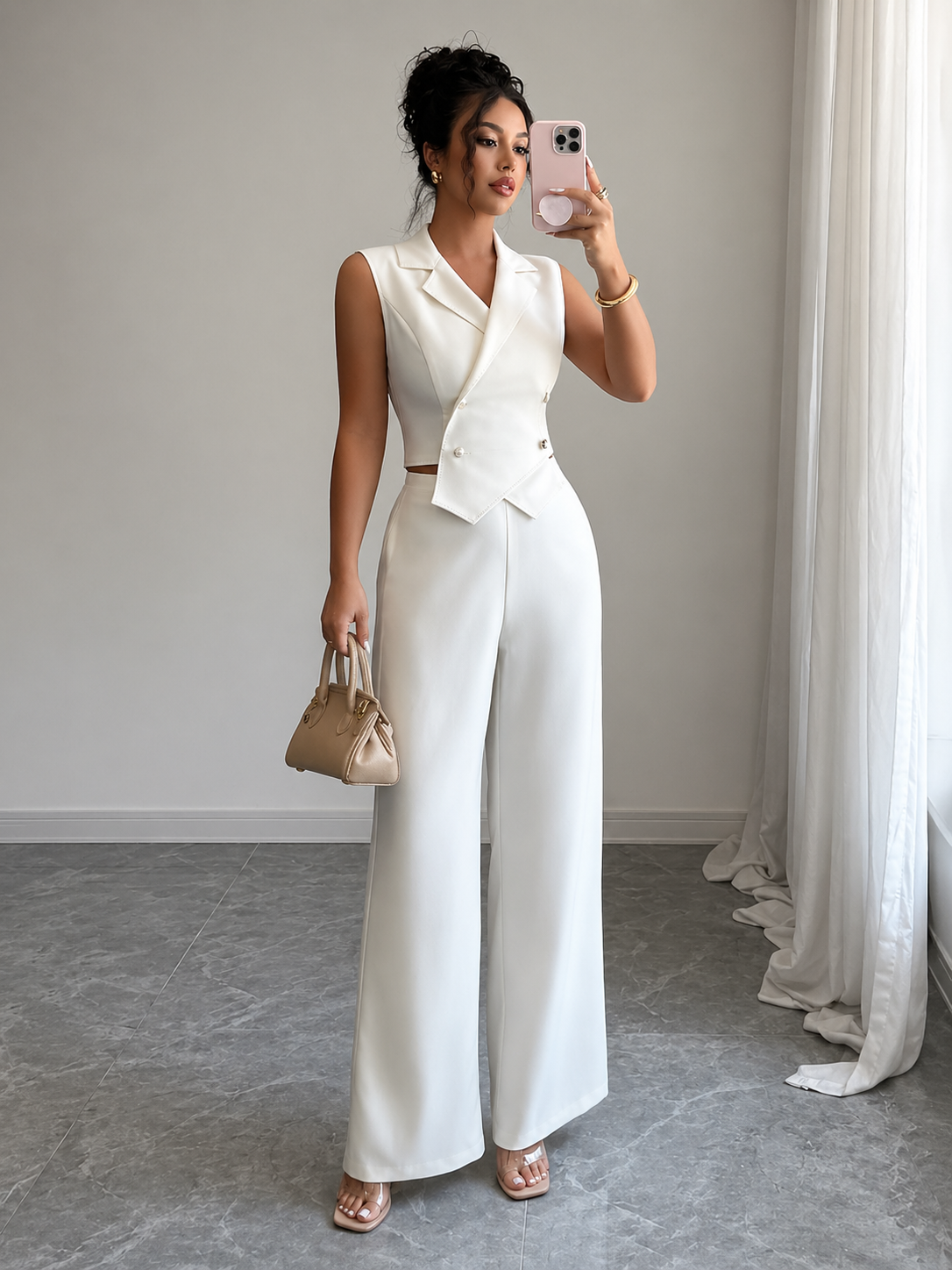 Woman in a white outfit taking a mirror selfie in a room with light gray walls and white curtains.