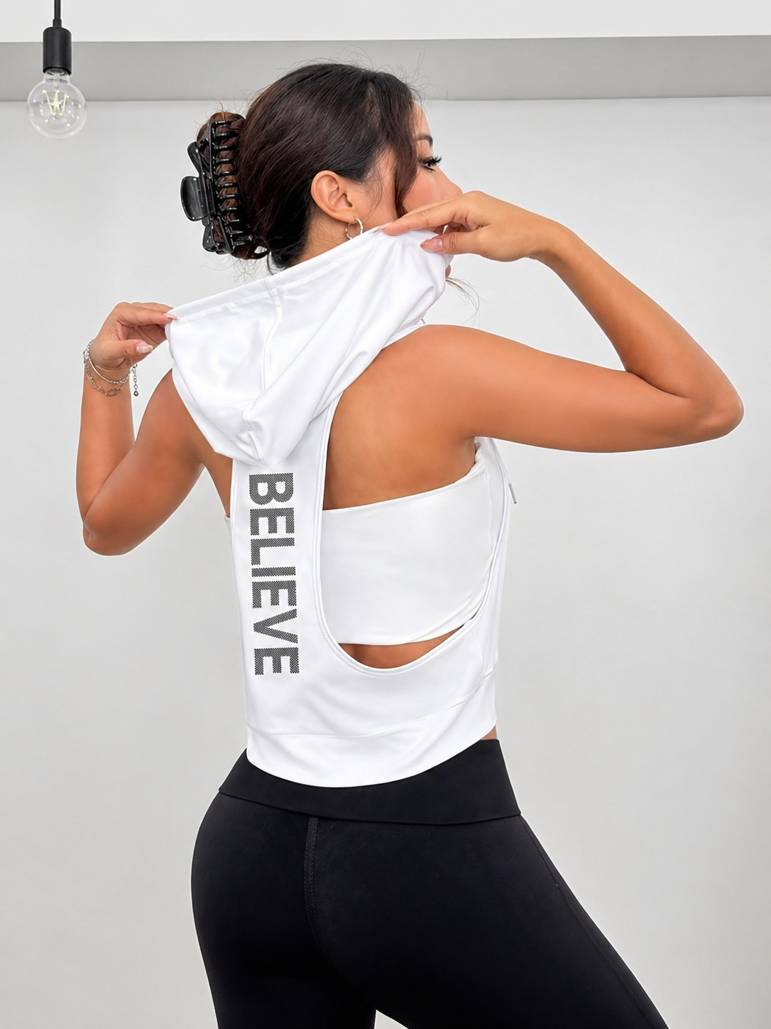 Woman wearing a white tank top with 'BELIEVE' text on the back, standing against a plain background.