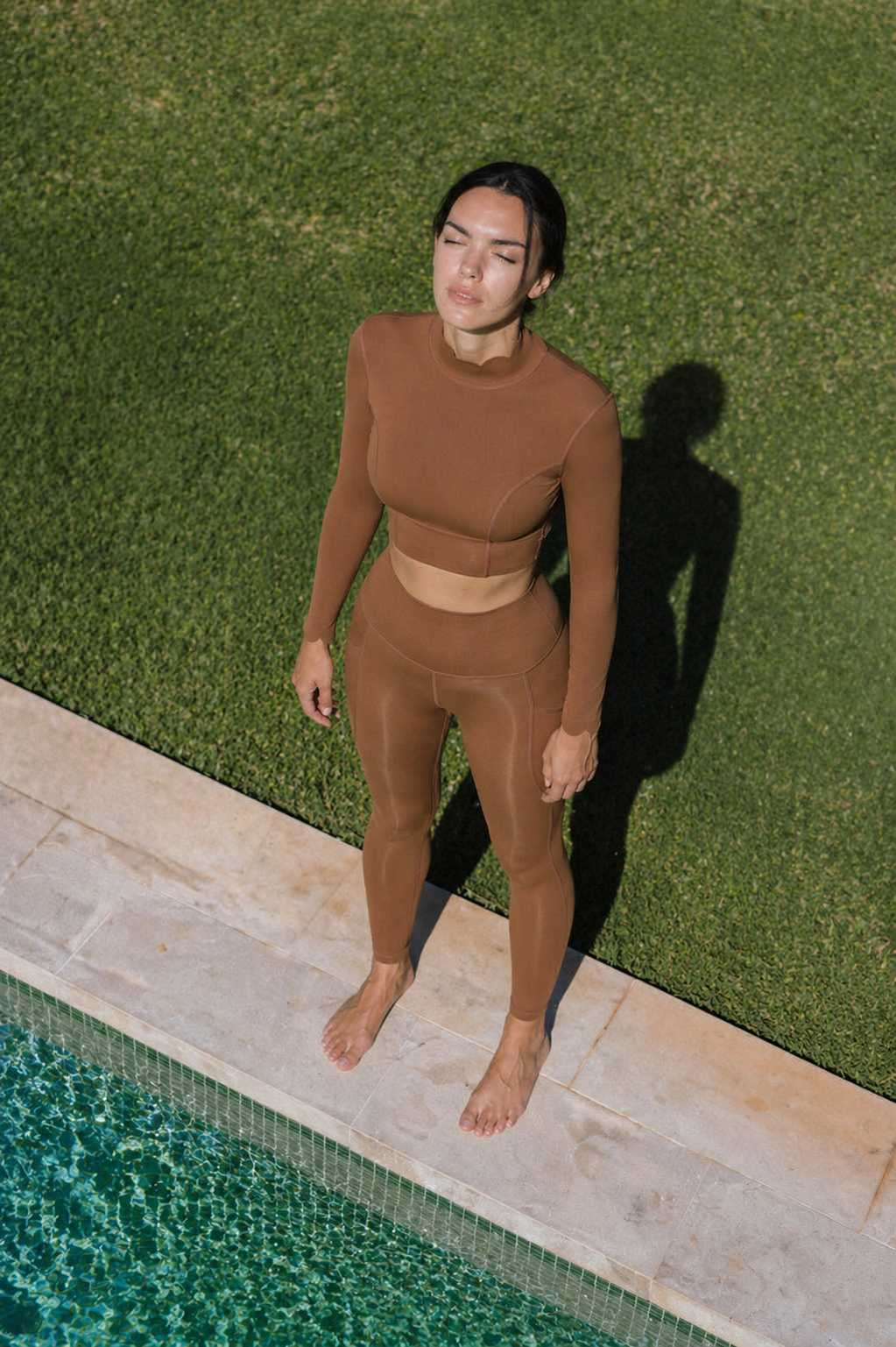 Person wearing a brown outfit standing by a pool with greenery in the background
