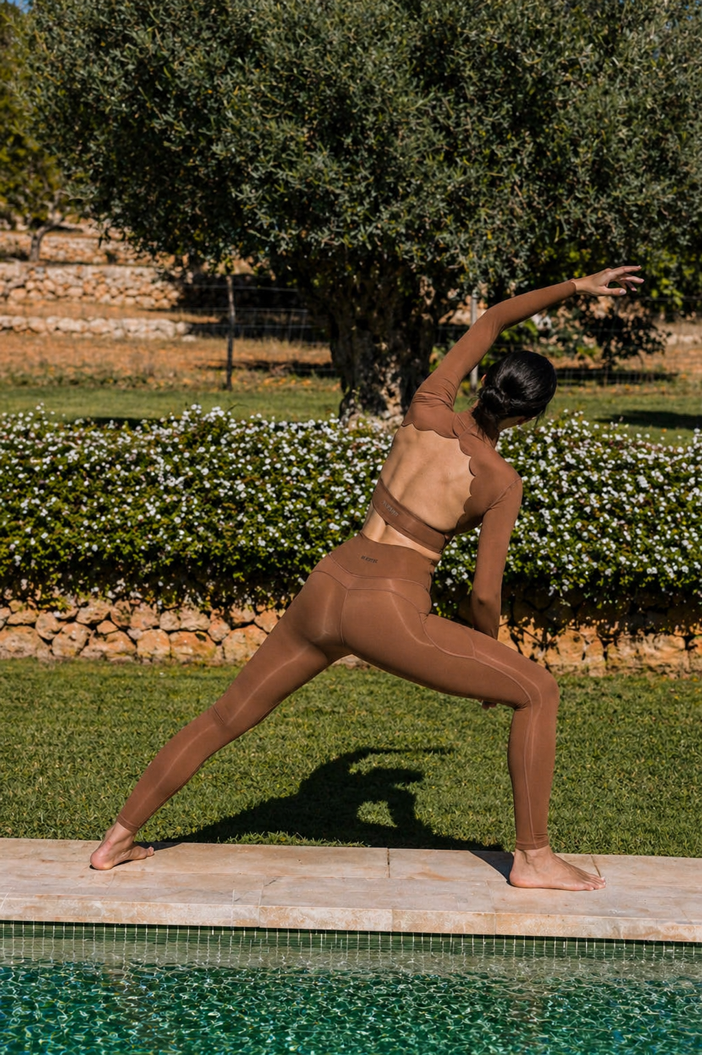 Person in brown athletic wear performing a yoga pose by a pool with greenery in the background