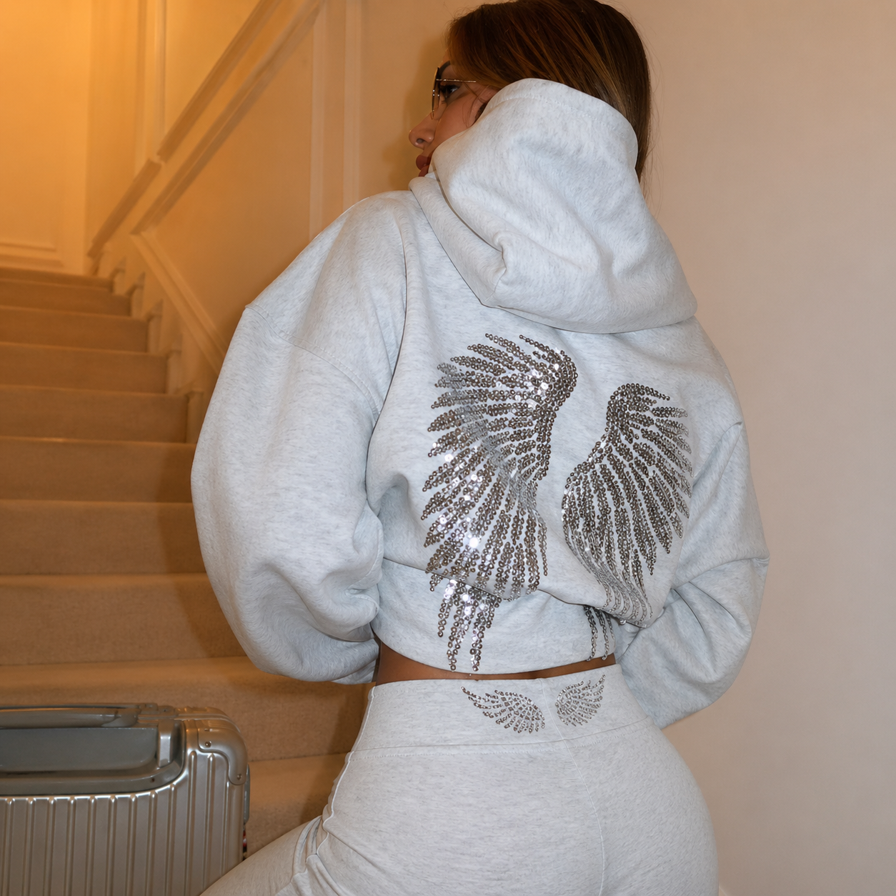 Person wearing a light gray hoodie with decorative wings on the back, standing in a room.