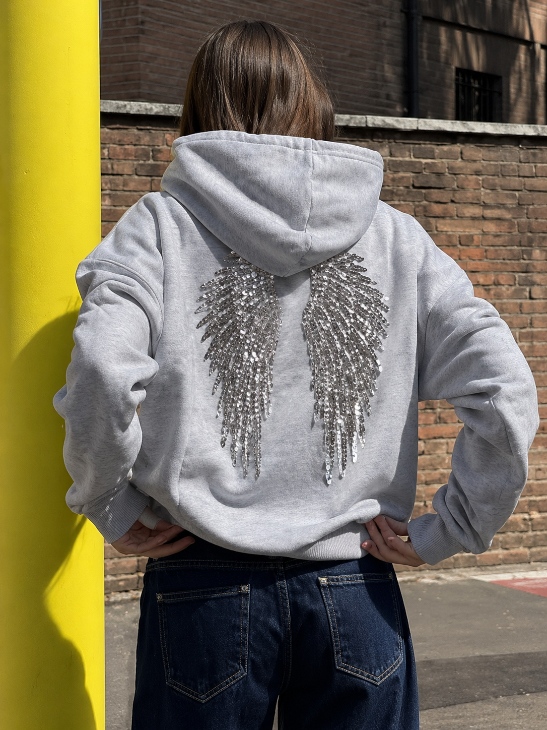 Person wearing a gray hoodie with sequin wings on the back, standing against a yellow wall and brick wall.
