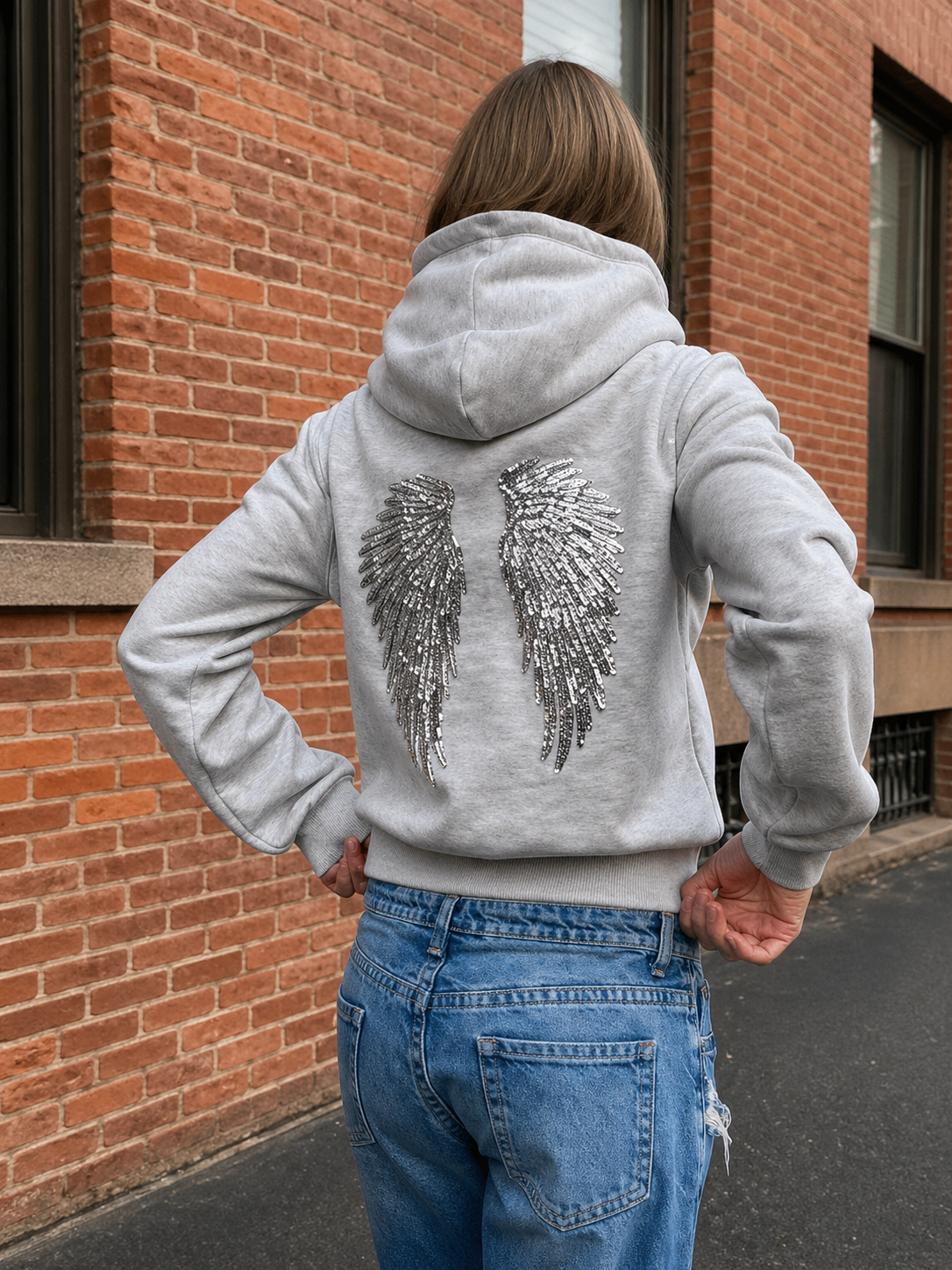 Person wearing a gray hoodie with sequin wings on the back, standing in front of a brick building.