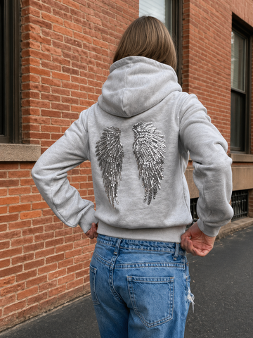 Person wearing a gray hoodie with sequin wings on the back, standing in front of a brick building.