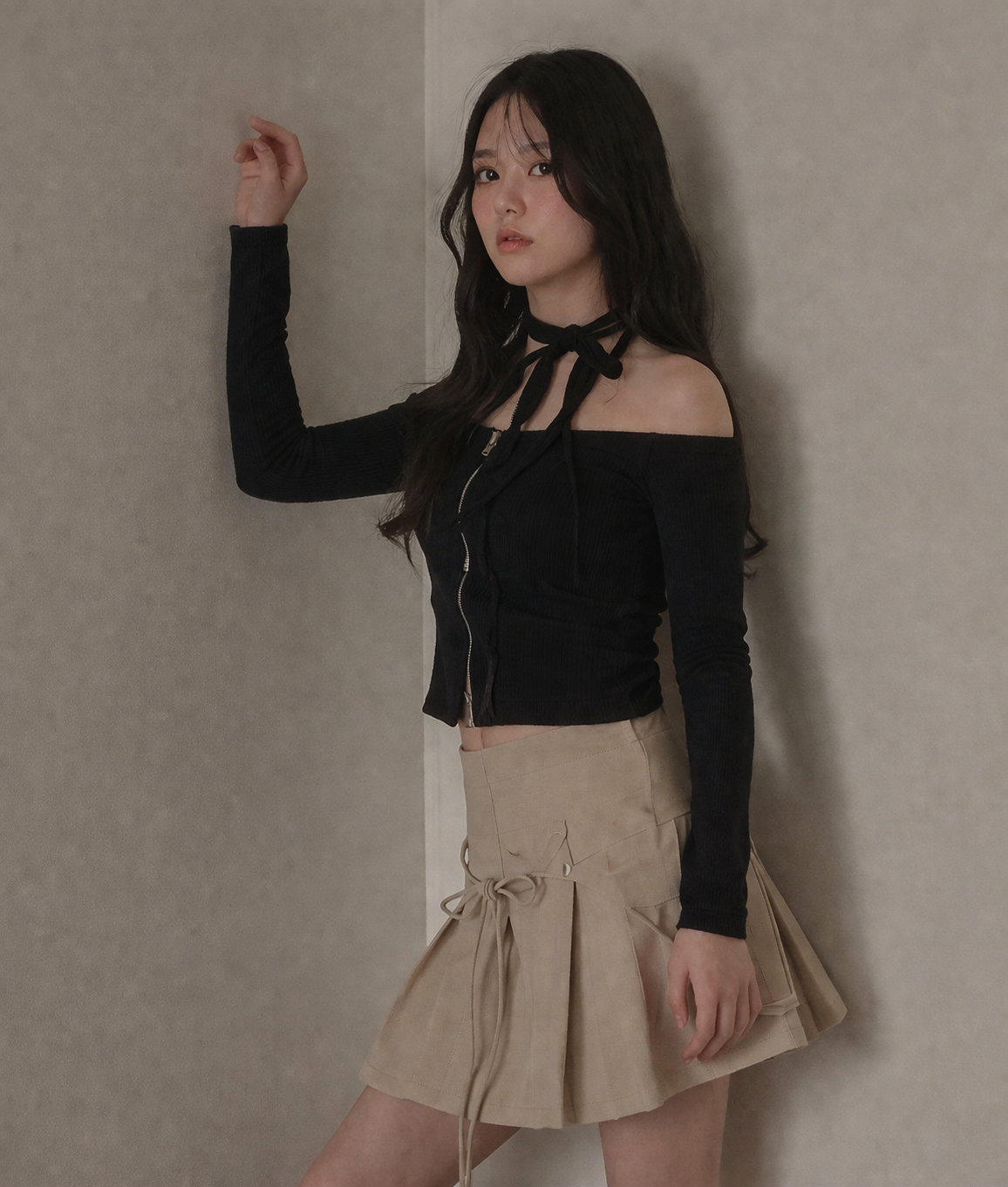 Woman wearing a black off-shoulder top and beige skirt against a plain wall.