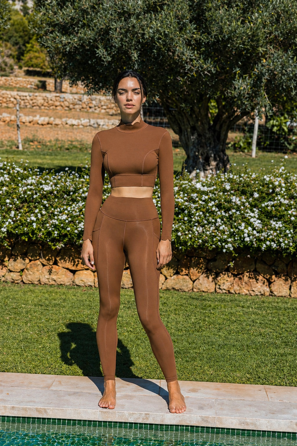 Woman in a brown outfit standing by a pool with greenery in the background